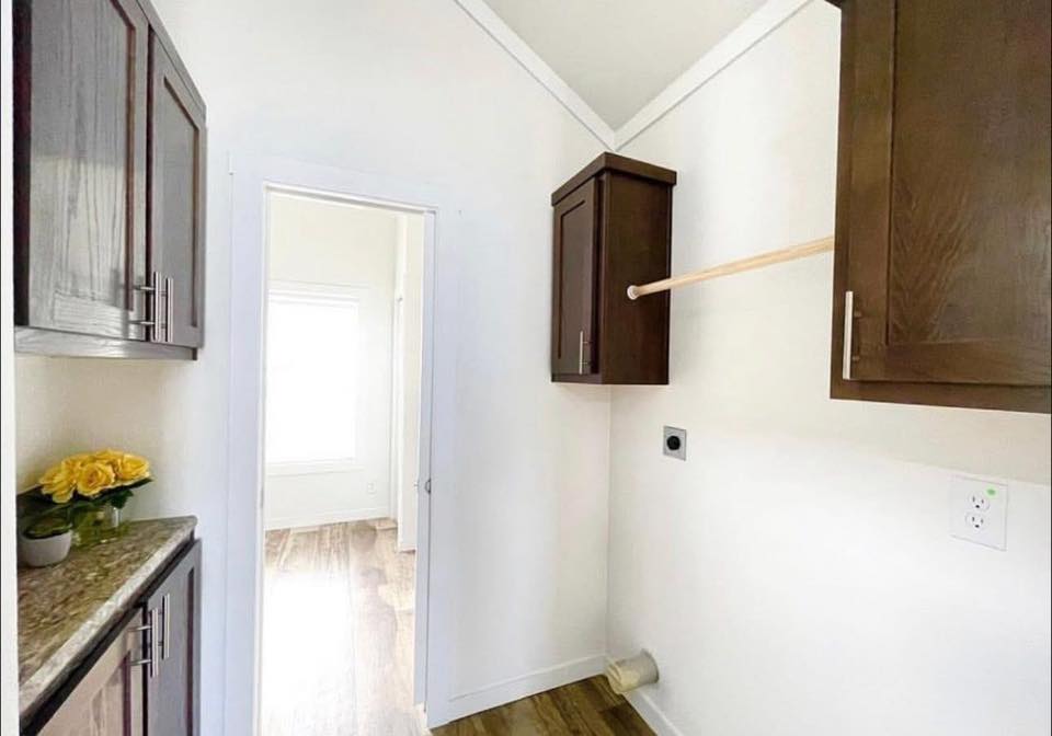 Narrow laundry room with dark wood cabinets, beige walls, and wood flooring. A hallway with sunlight is visible. Small flowers decorate the counter.