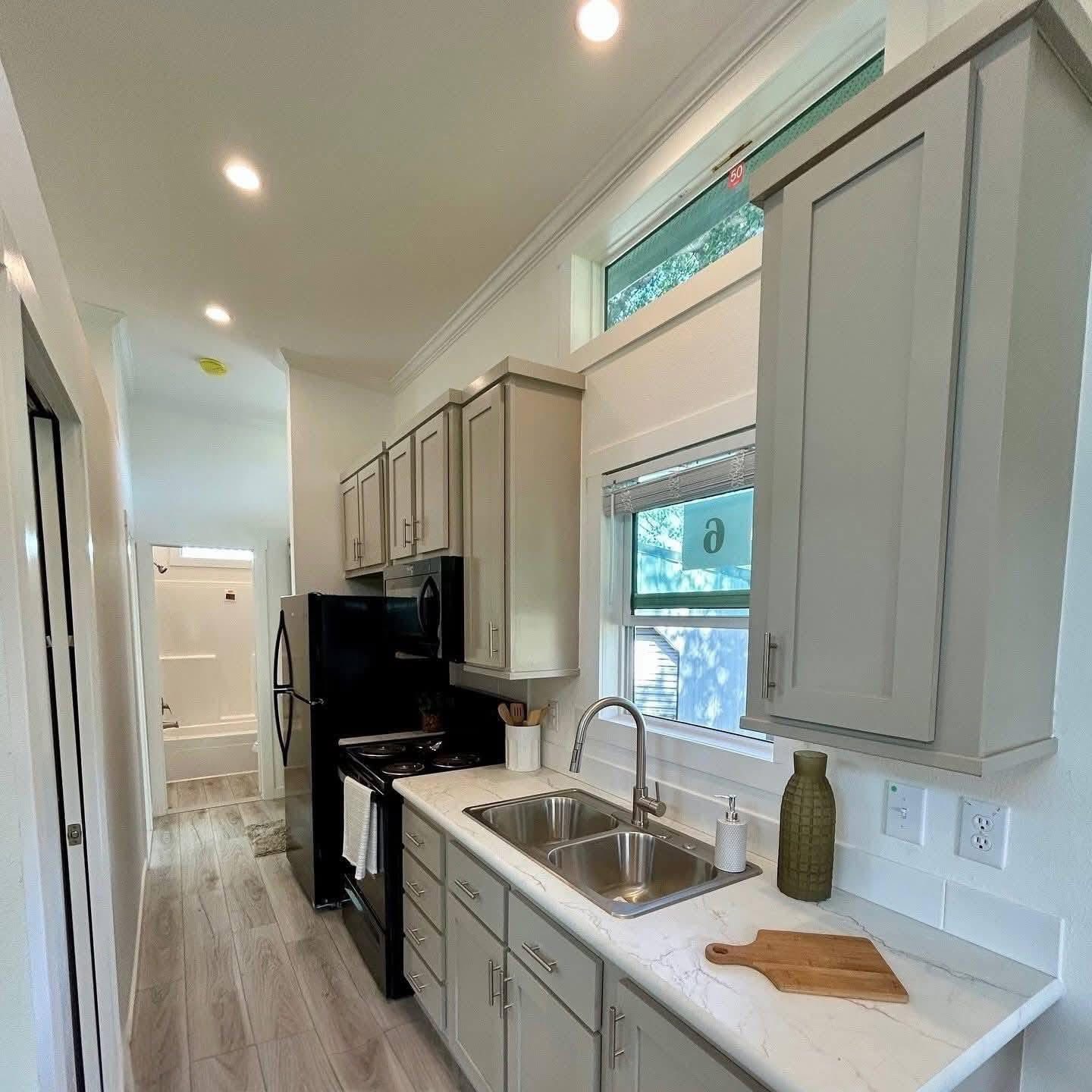 Narrow kitchen with grey cabinets, stainless steel appliances, and a marble countertop. Bright natural light from a window creates a fresh, modern feel.