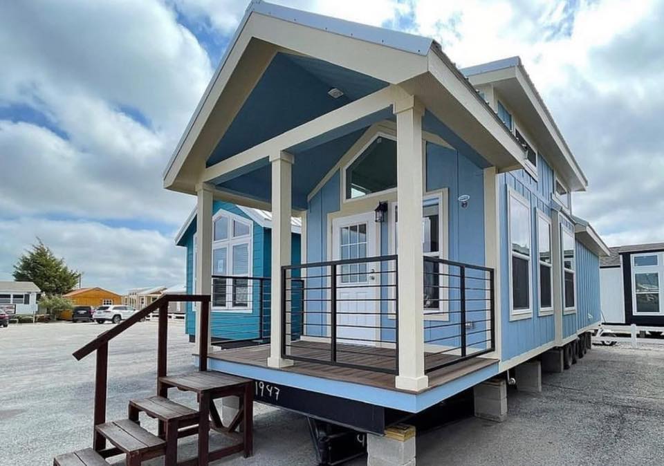 Small blue prefab home with a white-trimmed porch, wooden steps, and large windows. It sits on a lot under a cloudy sky, conveying a peaceful vibe.