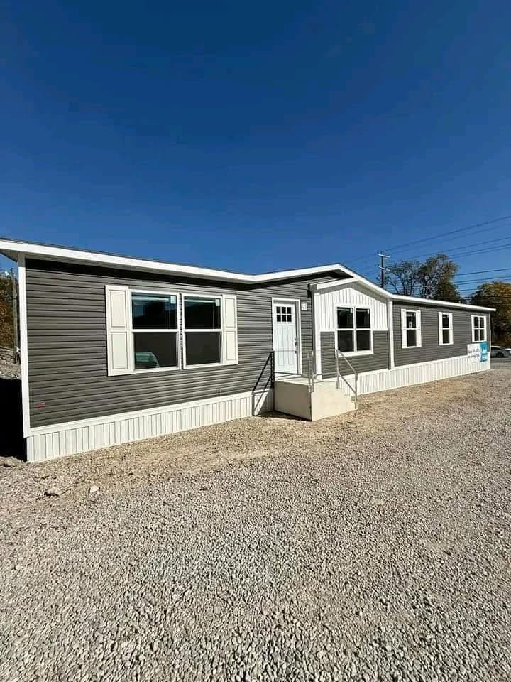 A modern, single-story manufactured home with gray siding and white trim under a clear blue sky. It features a small entry porch and multiple windows.