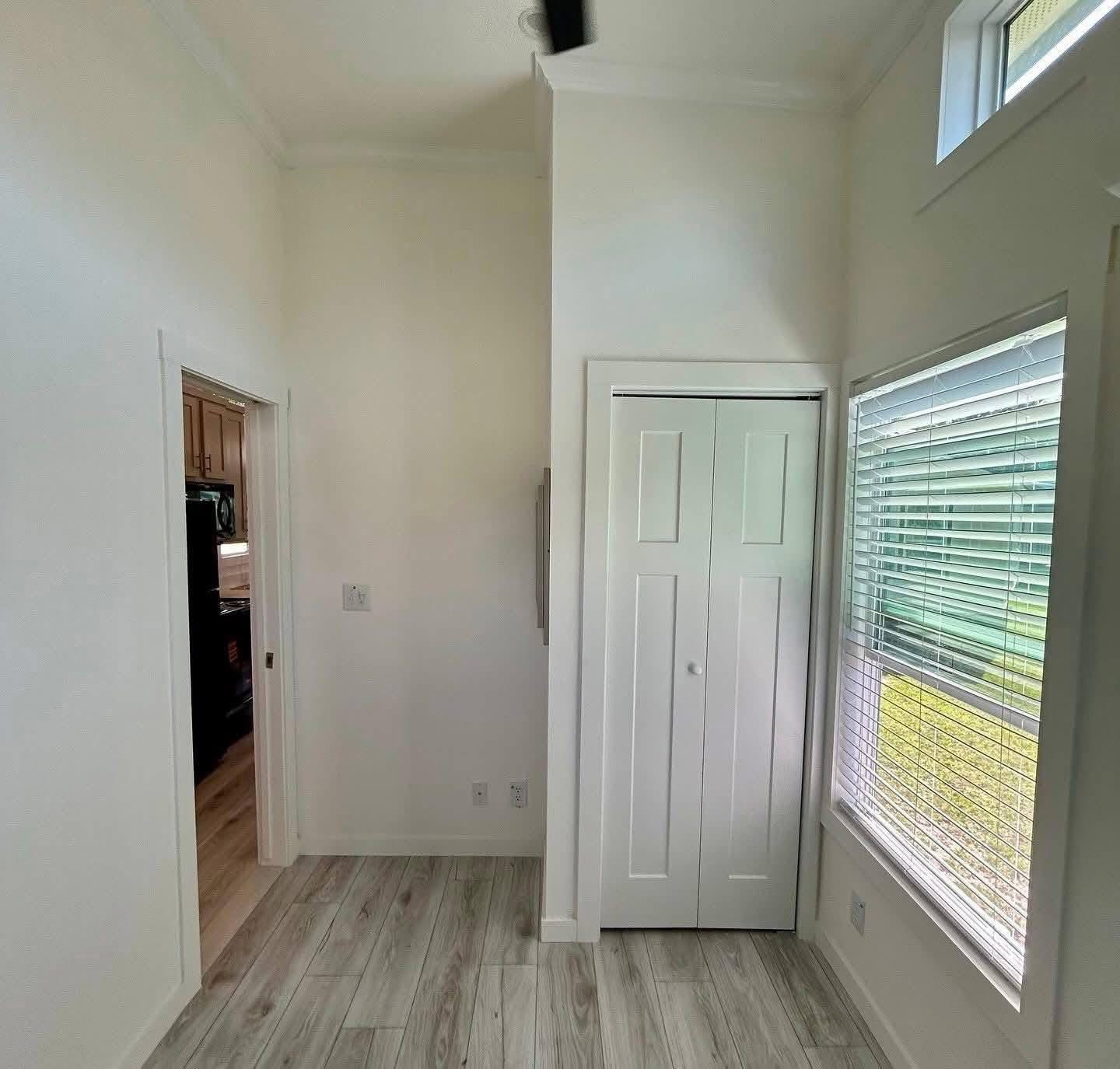 A small, bright room with white walls, light wood flooring, a narrow closet, and a large window with blinds. Sunlight creates a cozy atmosphere.