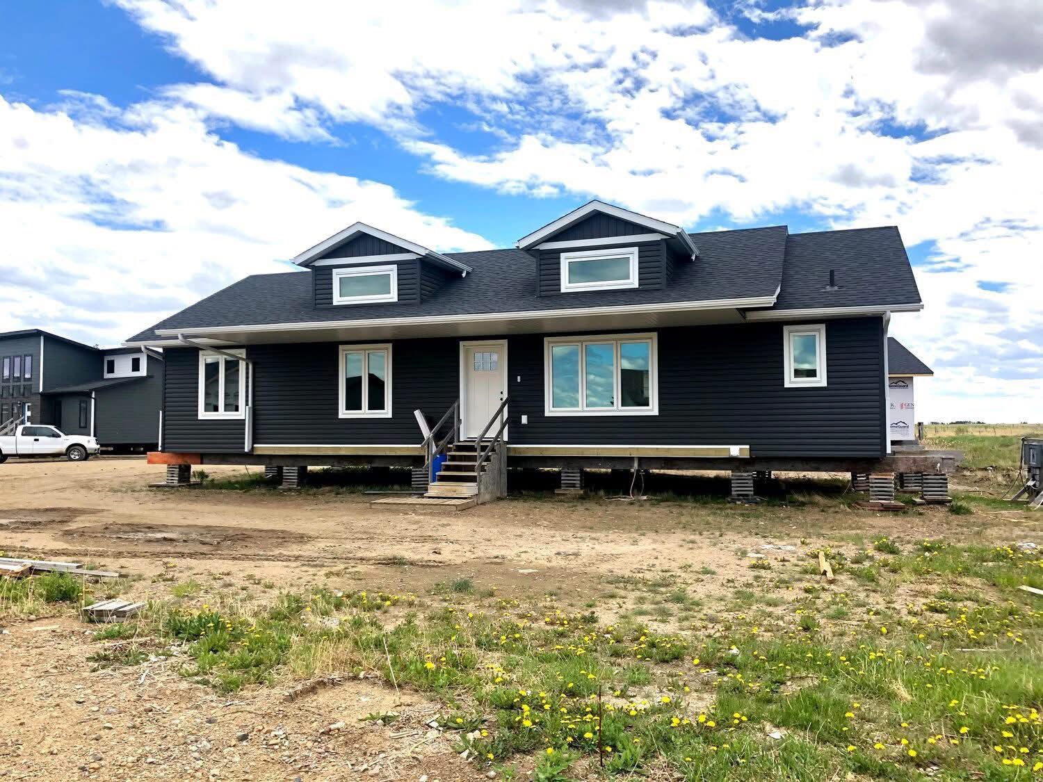 A dark gray, raised house with two dormer windows sits on a dirt lot under a cloudy blue sky. A white car and another house are visible in the background.