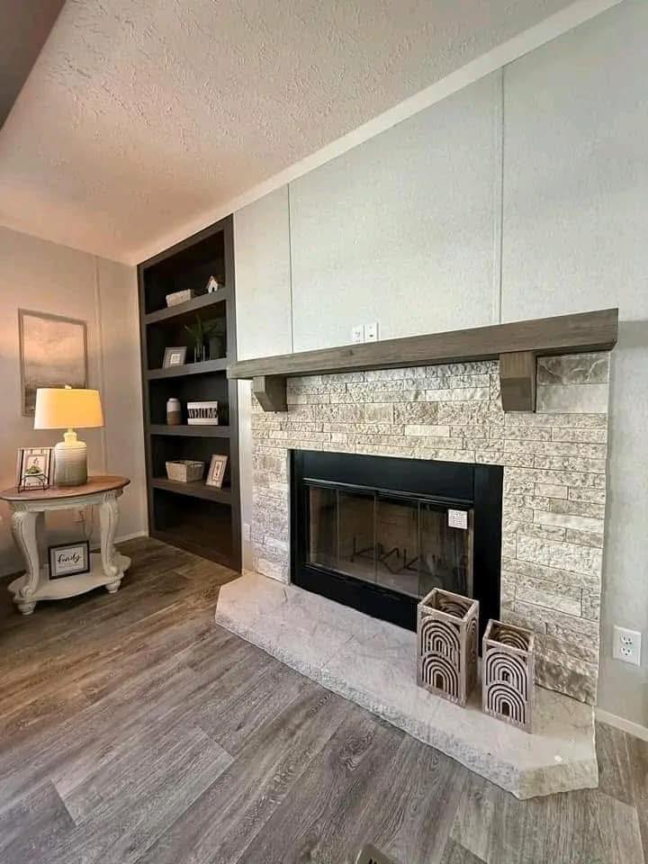 Cozy living room with stone fireplace, wooden mantel, and dark shelving. A side table with a lamp and decor elements adds warmth and elegance.