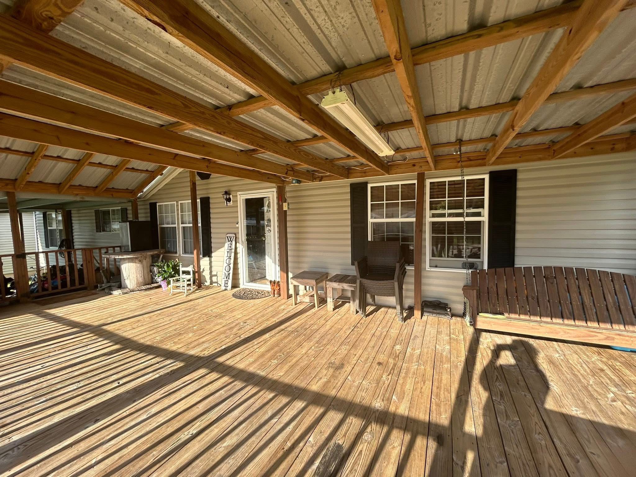 Spacious covered porch with wooden deck, featuring a swinging bench, chairs, and plants. Sunlight casts shadows, creating a warm, inviting atmosphere.