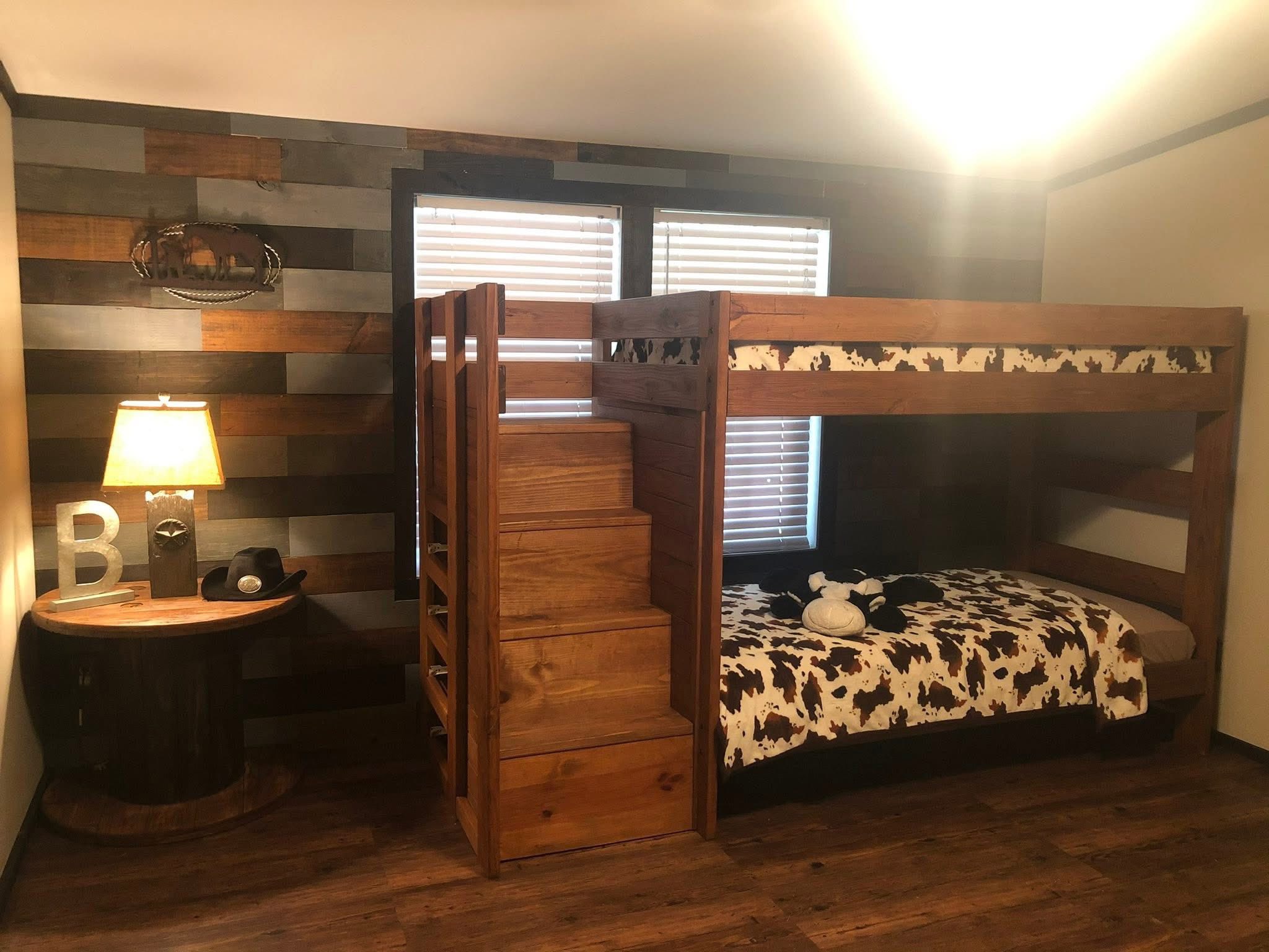 Cozy room with wooden bunk beds featuring a cow-print bedspread. A rustic wooden stairway leads to the top. A table with a lamp and decor is on the side.