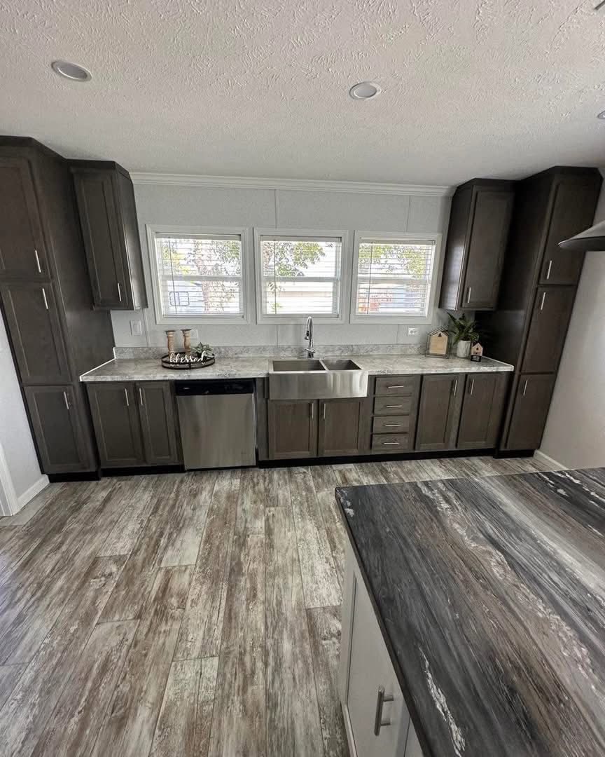 Spacious kitchen with dark wood cabinets, marbled countertops, stainless steel sink, and dishwasher. Bright light from three windows above.