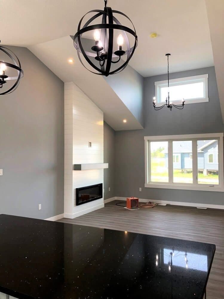 Spacious modern living room with high vaulted ceiling, two black chandeliers, large window, sleek fireplace, and glossy black countertop.