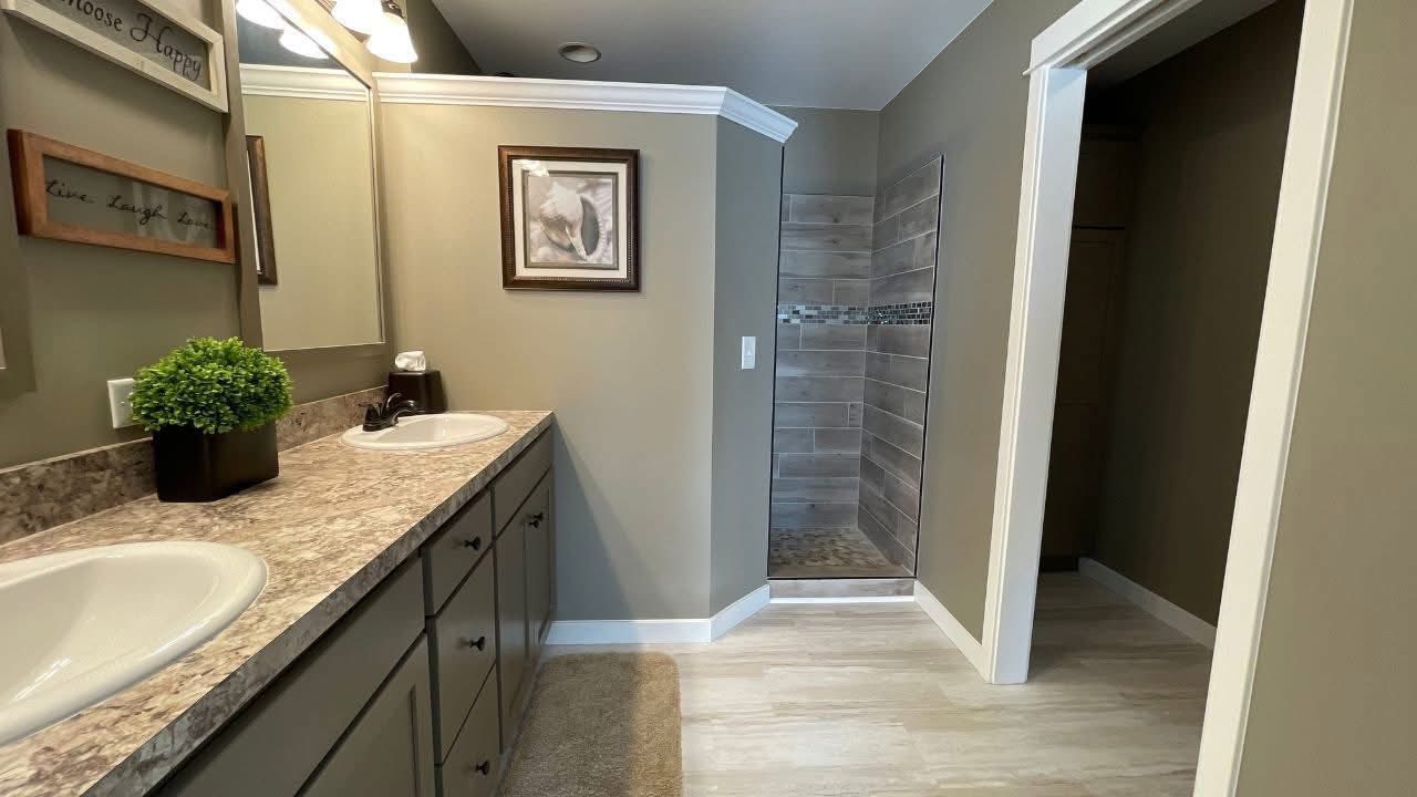 Modern bathroom with dual sinks, granite countertop, and wooden cabinets. A plant adds greenery. Tiled shower and framed art create a welcoming ambiance.