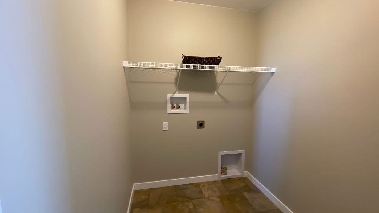 A minimalist laundry room with beige walls and tiled floor. A white wire shelf holds a wicker basket. Wall cutouts for a dryer and washer are visible.