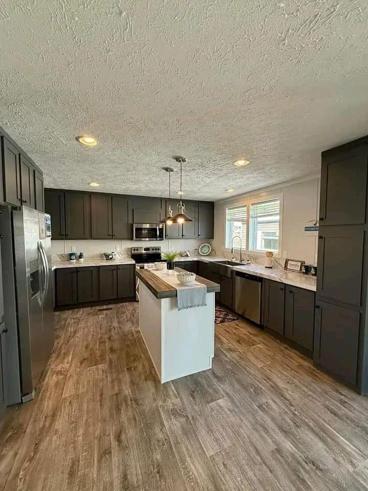Modern kitchen with dark cabinets, marble countertops, and stainless steel appliances. Large sink with a tall faucet, wooden floor, and bright overhead lighting.