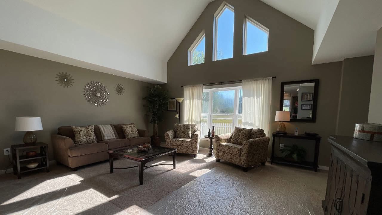 Spacious living room with high ceilings, a large triangular window, and sunlight streaming in. Neutral-toned furniture and decorative wall art create a cozy, inviting atmosphere.