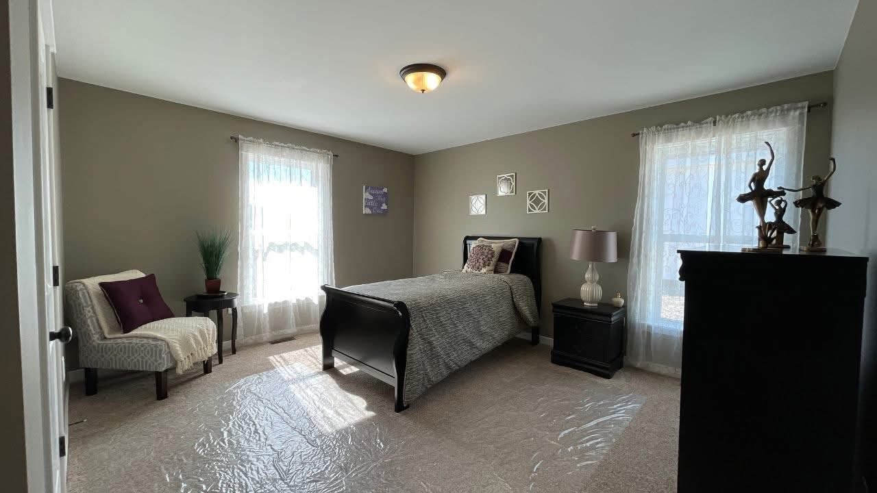 A cozy bedroom with a single bed, gray comforter, and decorative pillows. Two windows with sheer curtains allow light inside the room. A white armchair and small table with a plant sit to the left, while a side table with a lamp is on the bed's right. Wall art and a few sculptures provide a touch of elegance. The atmosphere is peaceful and inviting.