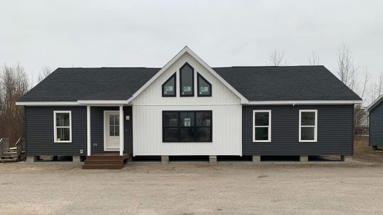 Single-story house with dark gray siding and white paneling. Features include a central gable with three windows, a dark roof, and small porch steps. Overcast day.