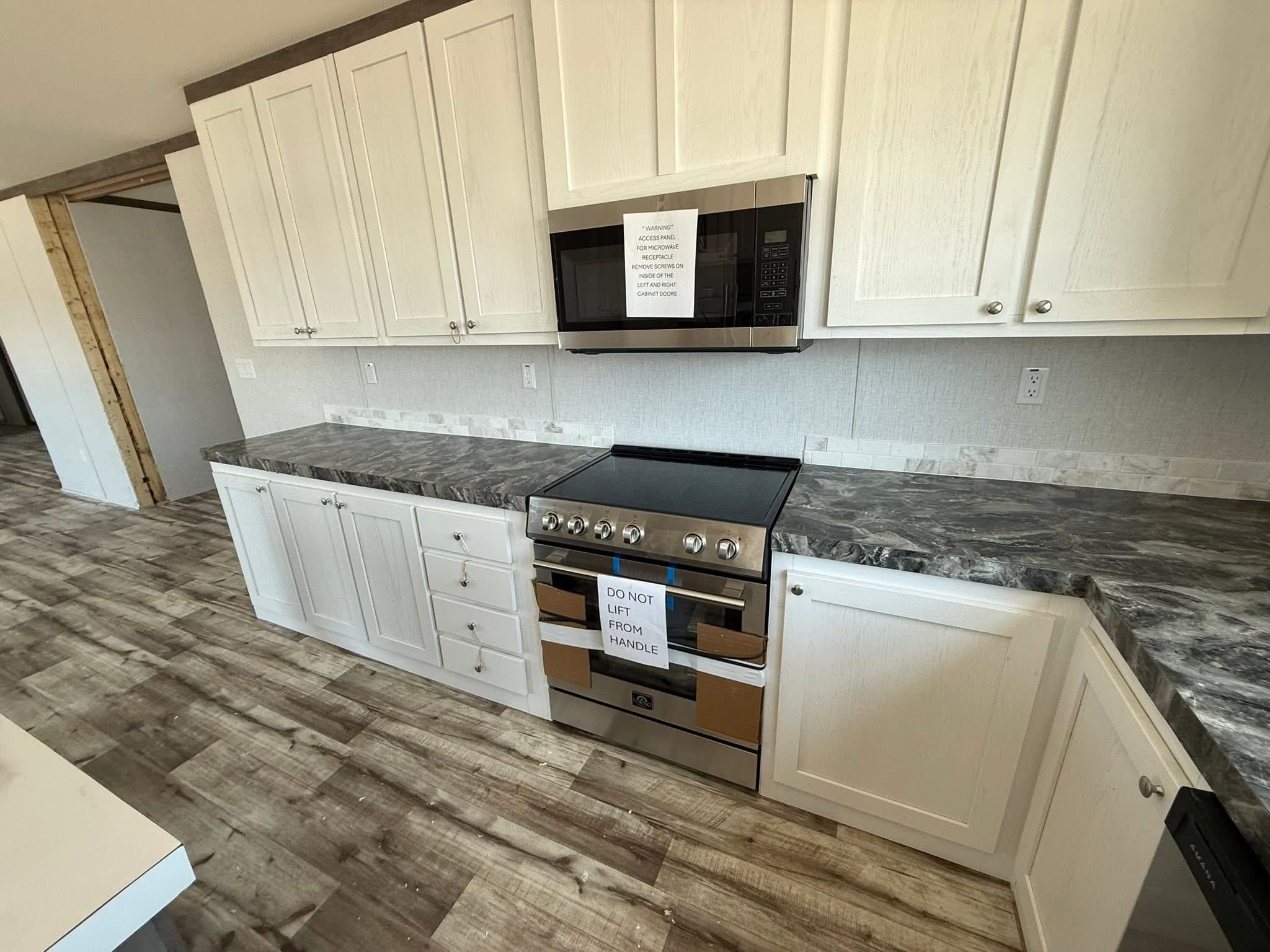 Modern kitchen with white cabinets, marble-patterned countertops, and stainless steel appliances. A note on the oven says, "Do not lift from handle." Neutral tones.