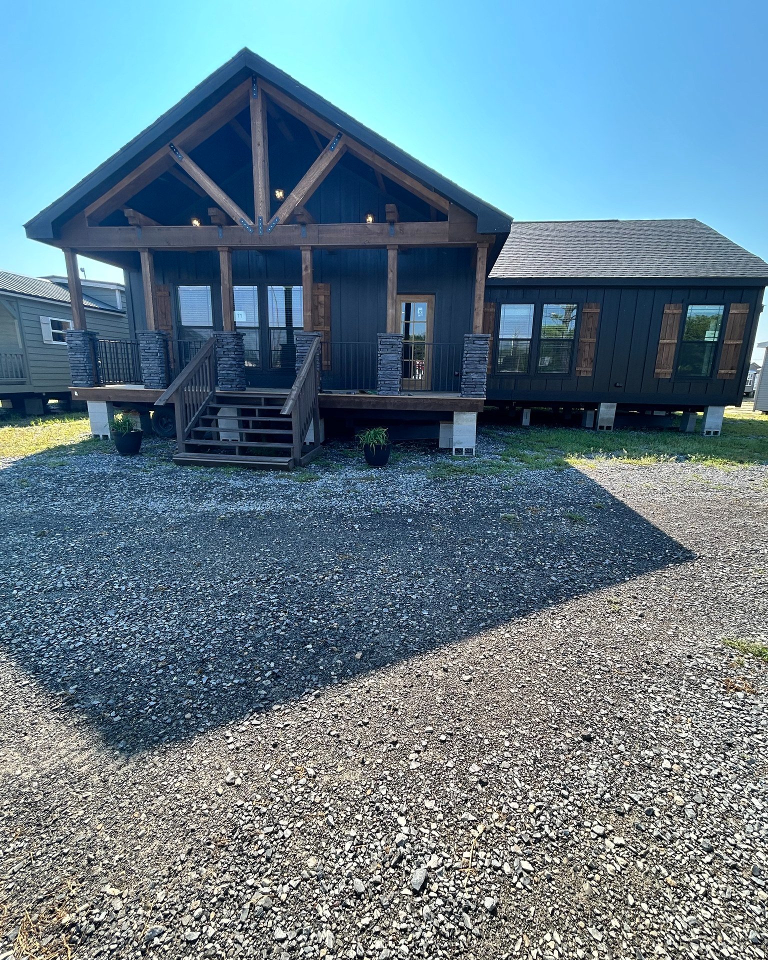 A modern cabin with a dark exterior, wooden accents, and a large porch with steps. Gravel driveway in front; blue sky above. Cozy and rustic feel.