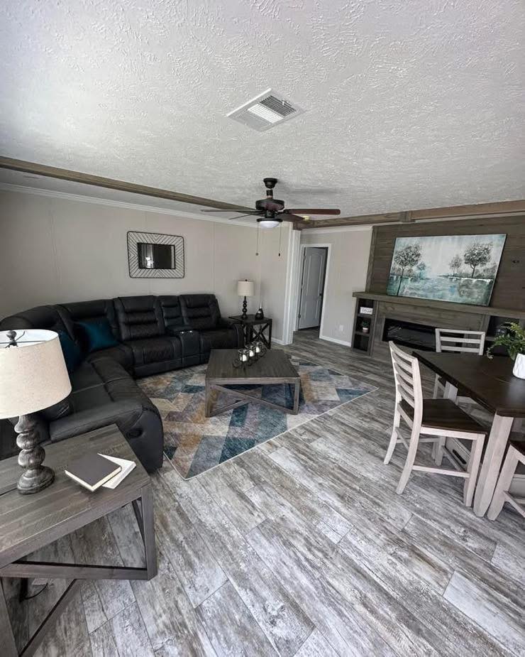 Modern living room with gray wood flooring, a black sectional sofa, geometric rug, rustic tables, and wall art. Bright and cozy atmosphere.