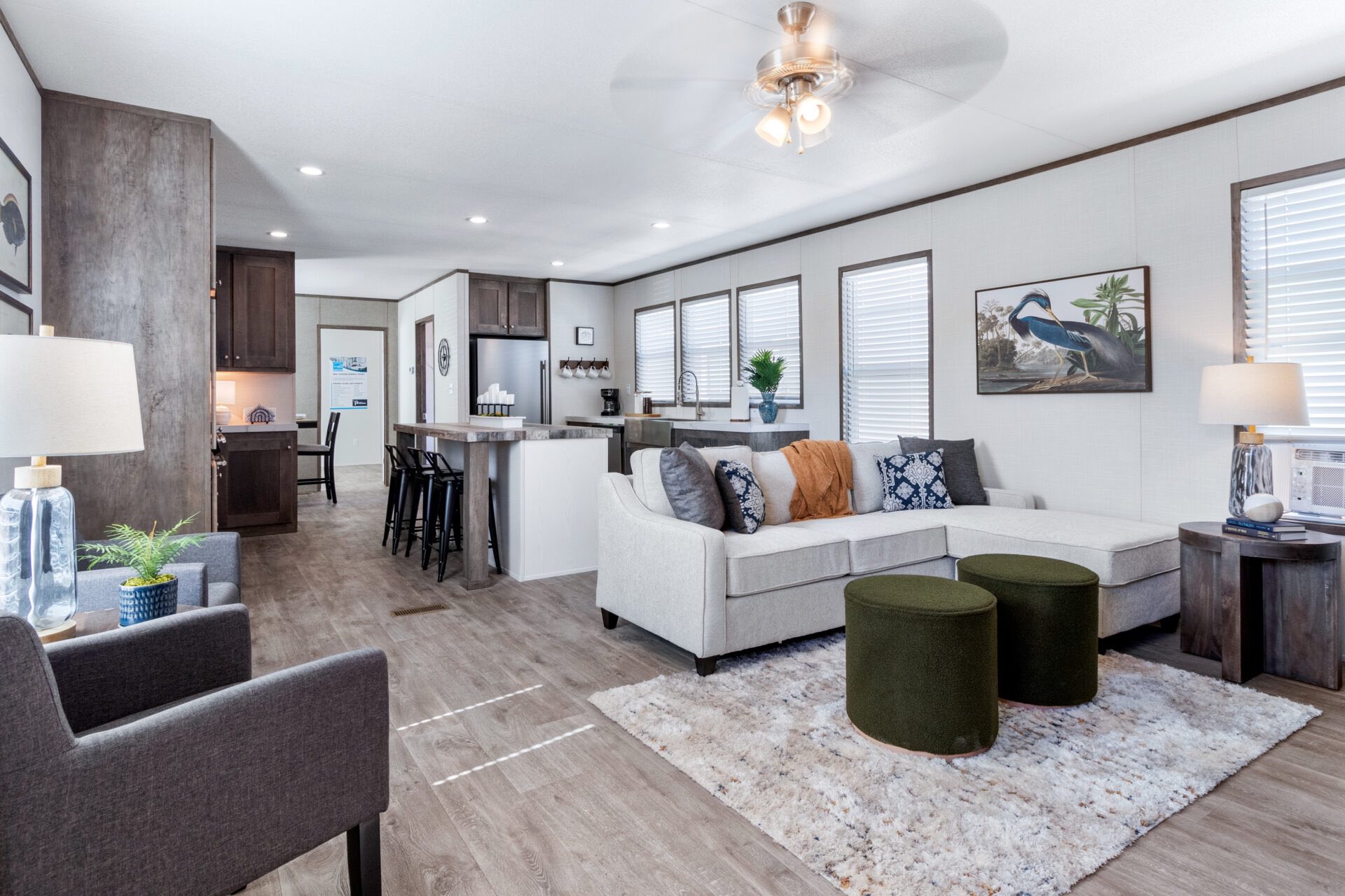 Modern open-plan living room with gray sectional sofa, green stools on a textured rug, and brown kitchen cabinets. The space feels cozy and bright.