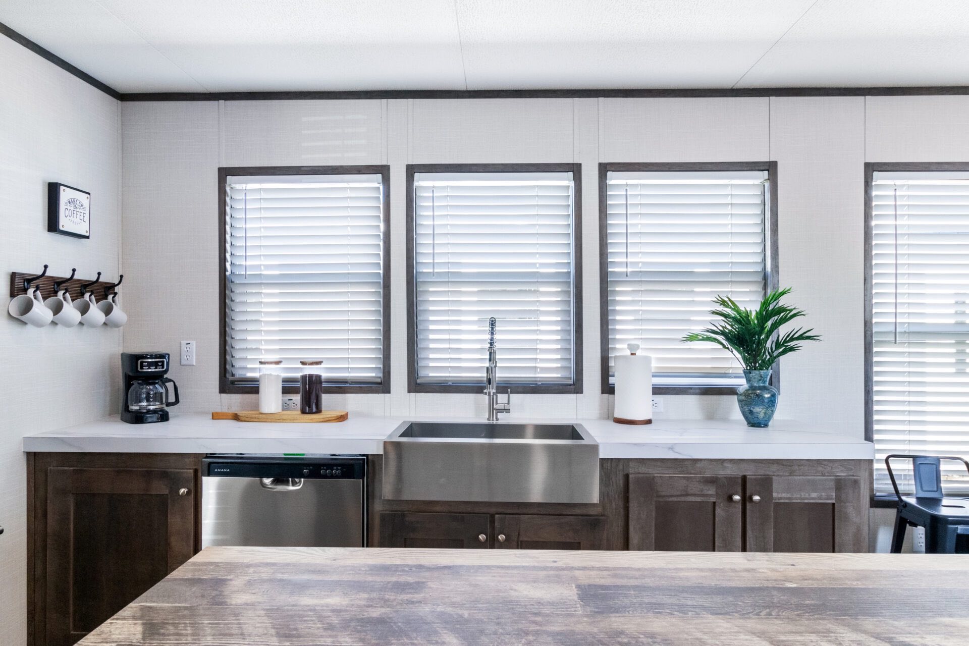 Modern kitchen with a large stainless steel sink, surrounded by white countertops and dark wood cabinets. Three windows with blinds allow soft light in. A coffee maker, potted plant, and kitchen essentials add warmth and functionality.