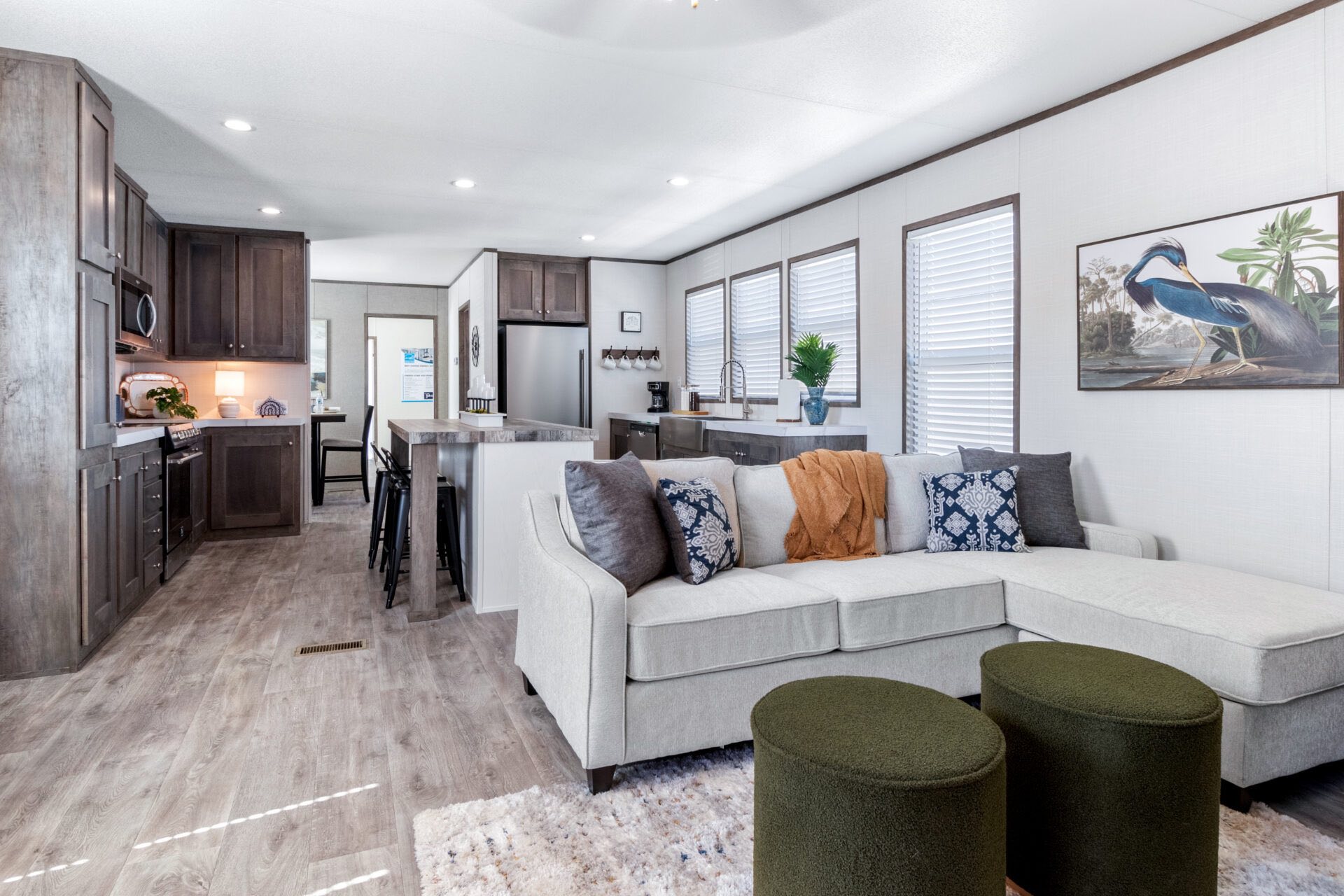 Open-concept living space with a modern kitchen and cozy living room. Features gray cabinets, a beige sectional sofa, colorful cushions, and wall art.