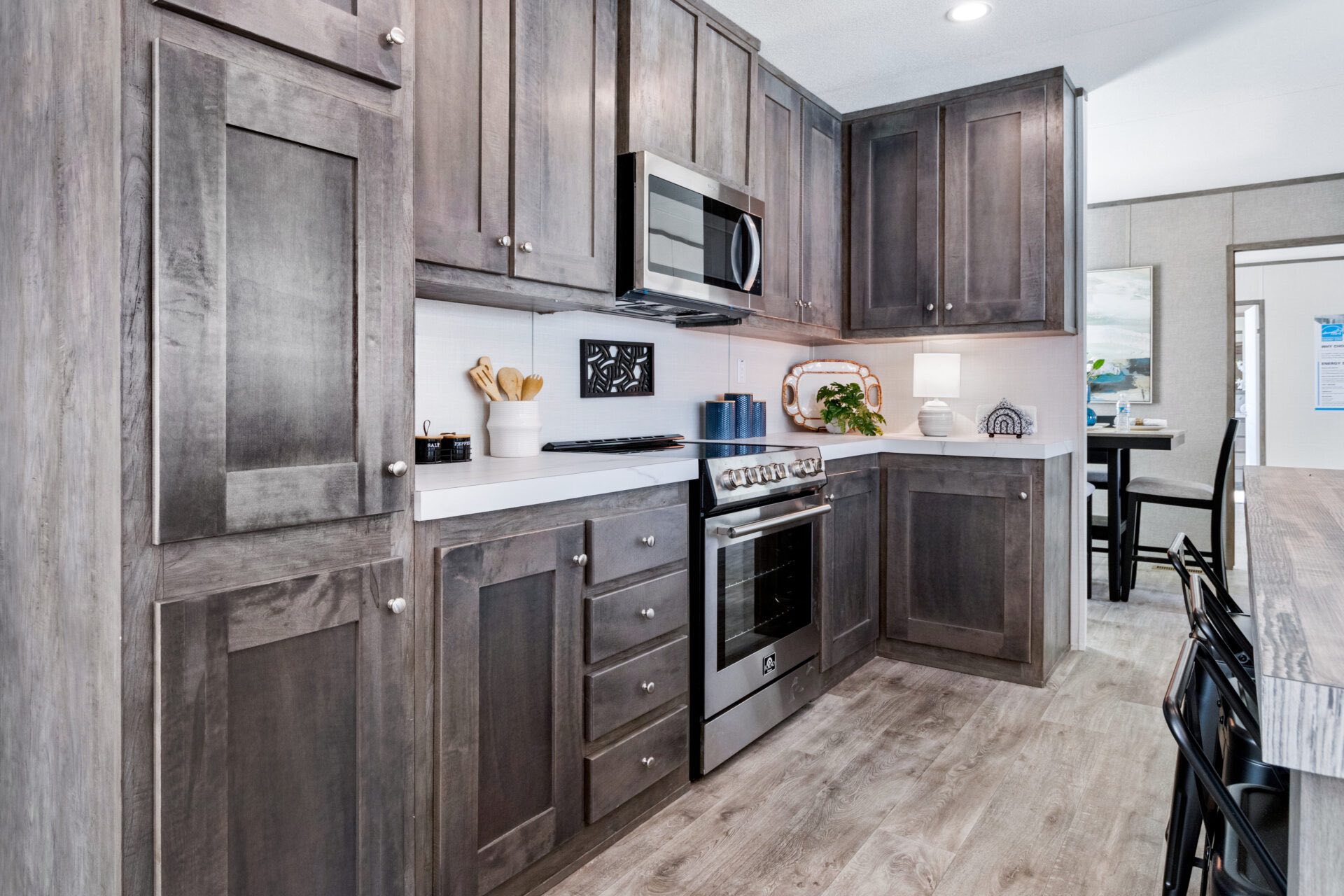 Modern kitchen with dark wooden cabinets, stainless steel appliances, and light wood flooring. Bright atmosphere with decorative plants and utensils.