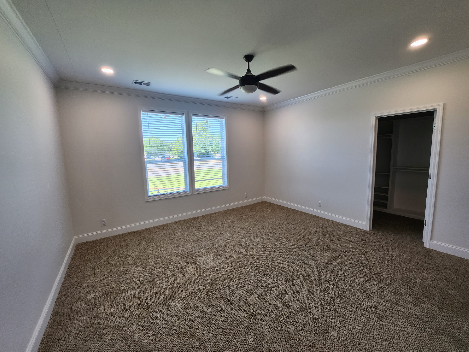 Spacious, empty bedroom with gray carpet, white walls, ceiling fan, large window, and an open walk-in closet. Natural light creates a calm, airy feel.