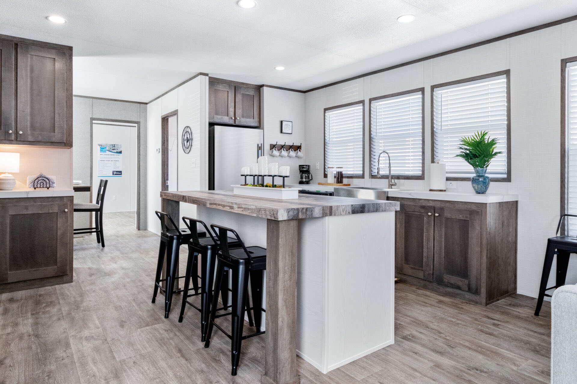 Bright kitchen with white walls and wood flooring. Features a central island with stools, dark wood cabinets, large windows, and modern appliances.