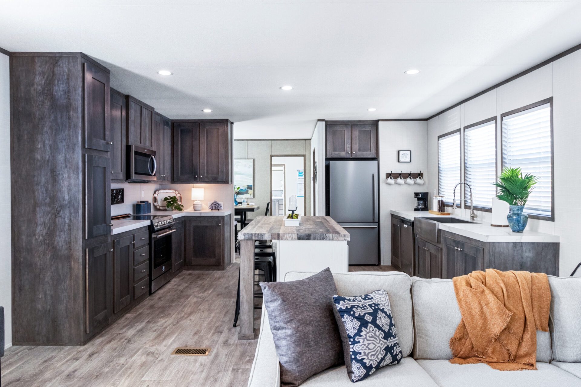 Modern kitchen and living space with dark wood cabinets, a central island, and sleek appliances. A cozy white sofa with colorful cushions and a brown throw adds warmth. Bright and inviting atmosphere.