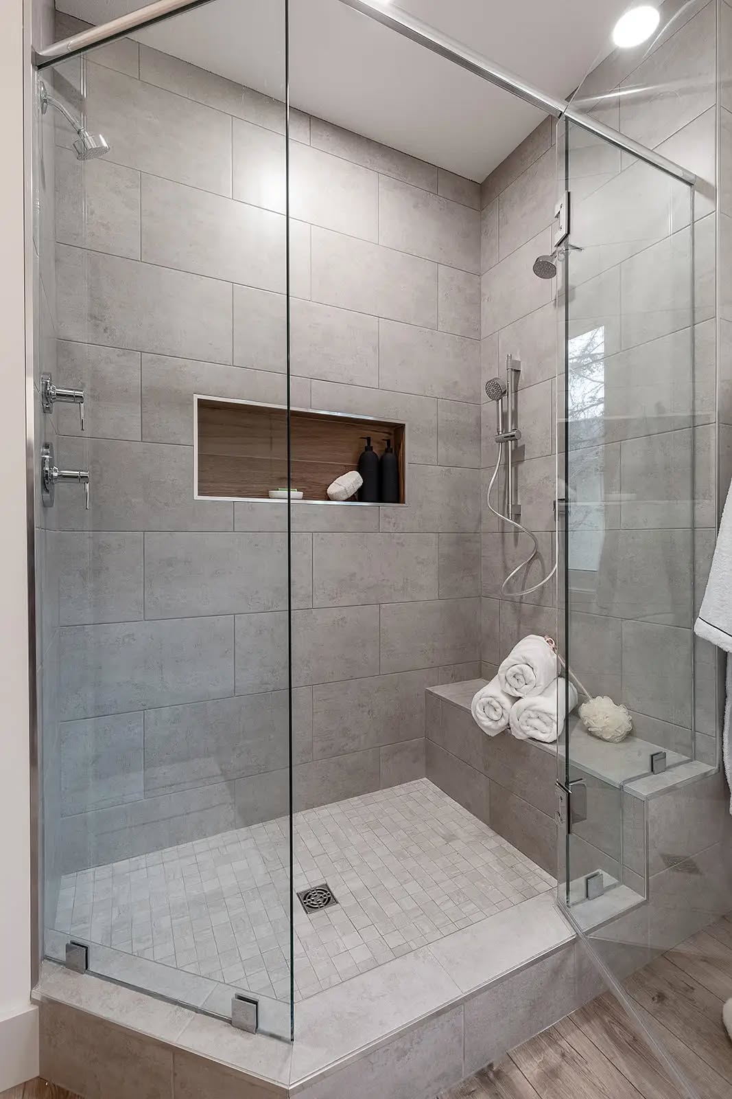 Modern shower with gray tiles and glass doors, featuring a built-in niche with dark bottles and towels. Soft lighting creates a clean, serene vibe.