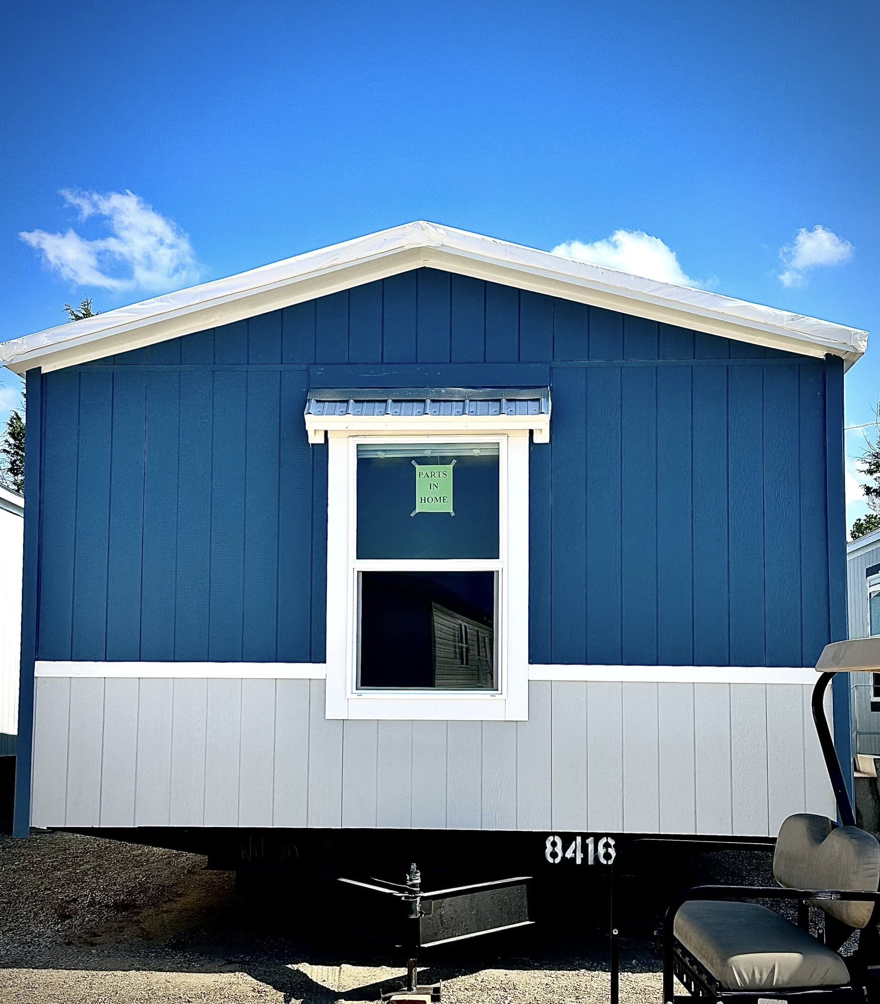 Blue and gray mobile home front against a bright blue sky with white clouds. A sign in the window reads "Paris in Home." A golf cart is partially visible on the right.
