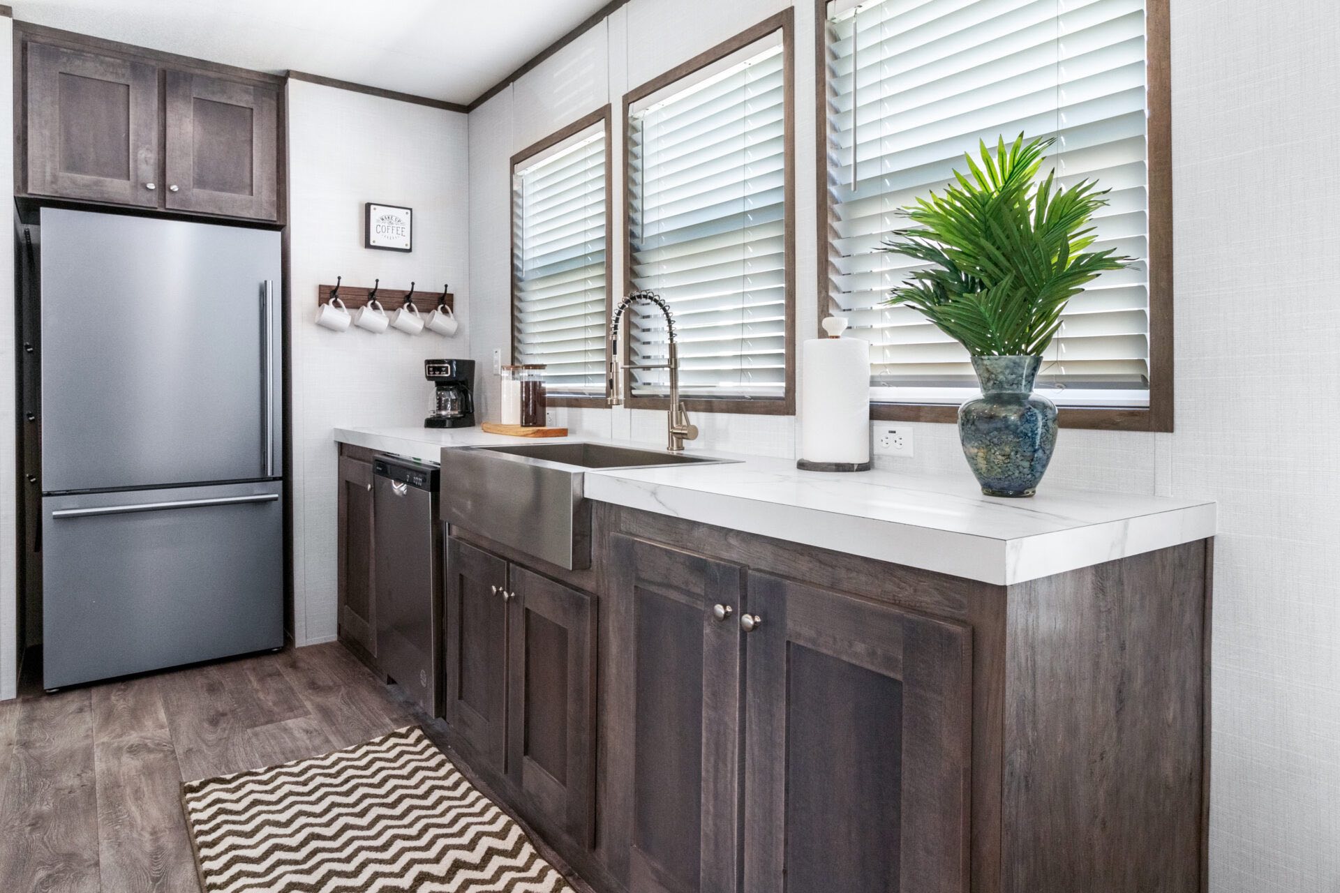 Modern kitchen with dark wood cabinets, stainless steel fridge, and a farmhouse sink. Countertop features a plant, paper towels, and coffee setup.