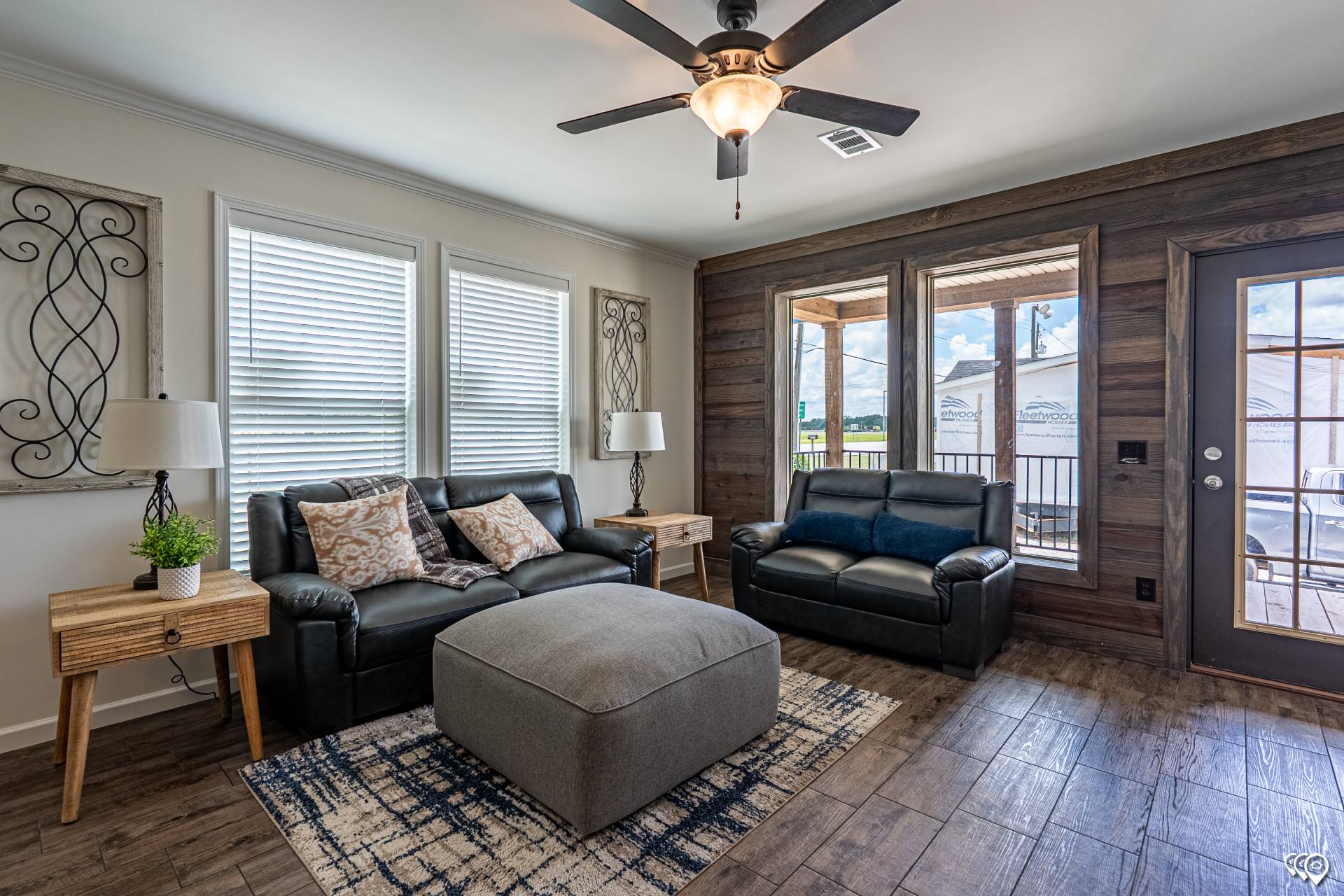 Cozy living room with leather sofas, a large ottoman, wooden side tables, and abstract wall decor. Ceiling fan spins above, windows brighten the space.