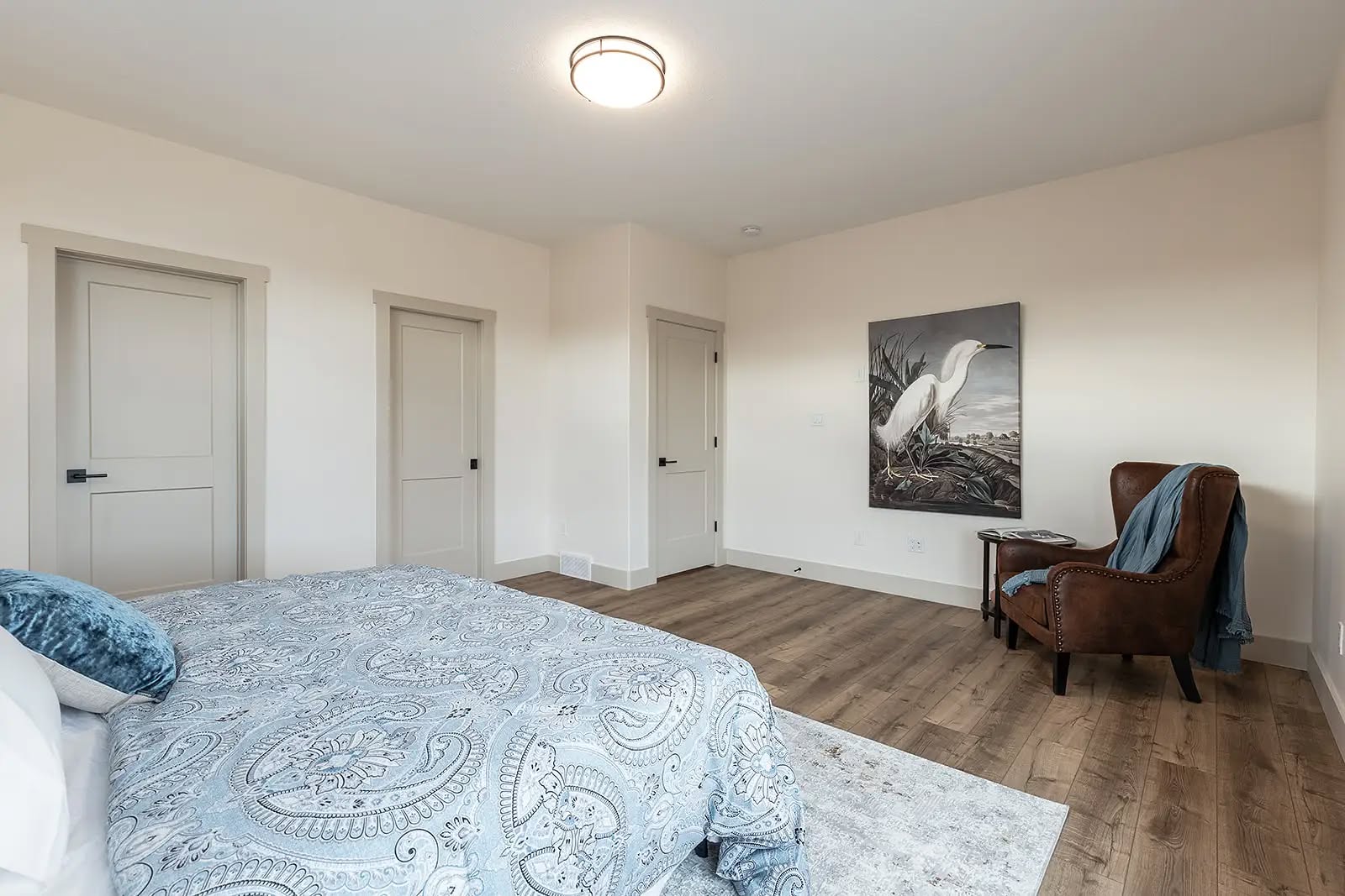 Spacious bedroom with light walls and hardwood floors. Features a bed with paisley bedding, leather armchair, heron artwork, and modern ceiling light. Calm ambiance.