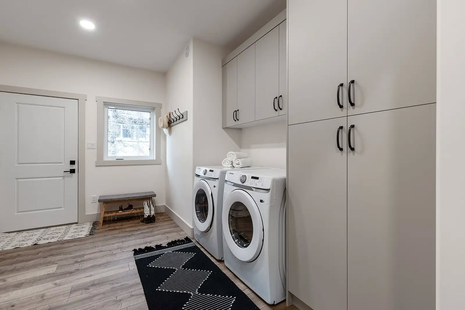 Modern laundry room with white walls and wood flooring. Features a front-loading washer and dryer, overhead cabinets, a small window, and a bench by the door.