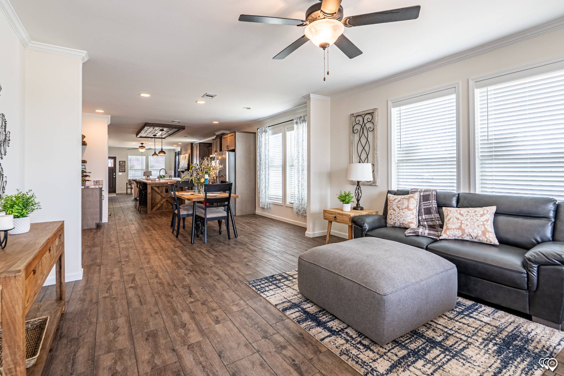 A spacious open-plan living room with hardwood floors and modern decor. It features a black sofa with patterned pillows, a large gray ottoman on a rug, and a dining area in the background. Sunlight streams through large windows, creating a cozy, inviting atmosphere.