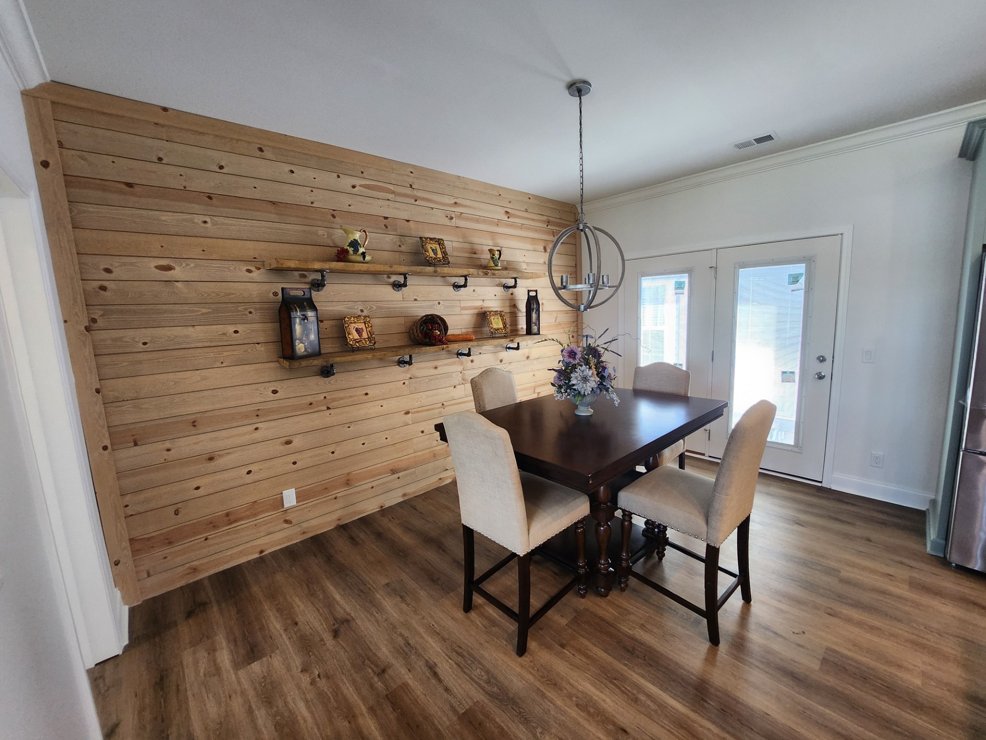 Dining room with wooden accent wall and floating shelves displaying decor. A dark wood table with beige chairs is under a modern chandelier. Bright, airy vibe.