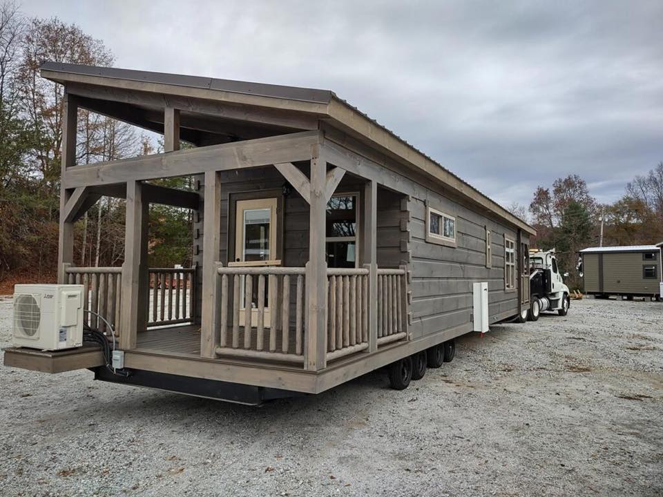 A rustic tiny house on wheels with wooden siding and a small porch, parked on gravel under a cloudy sky. A white truck is visible, ready for towing.