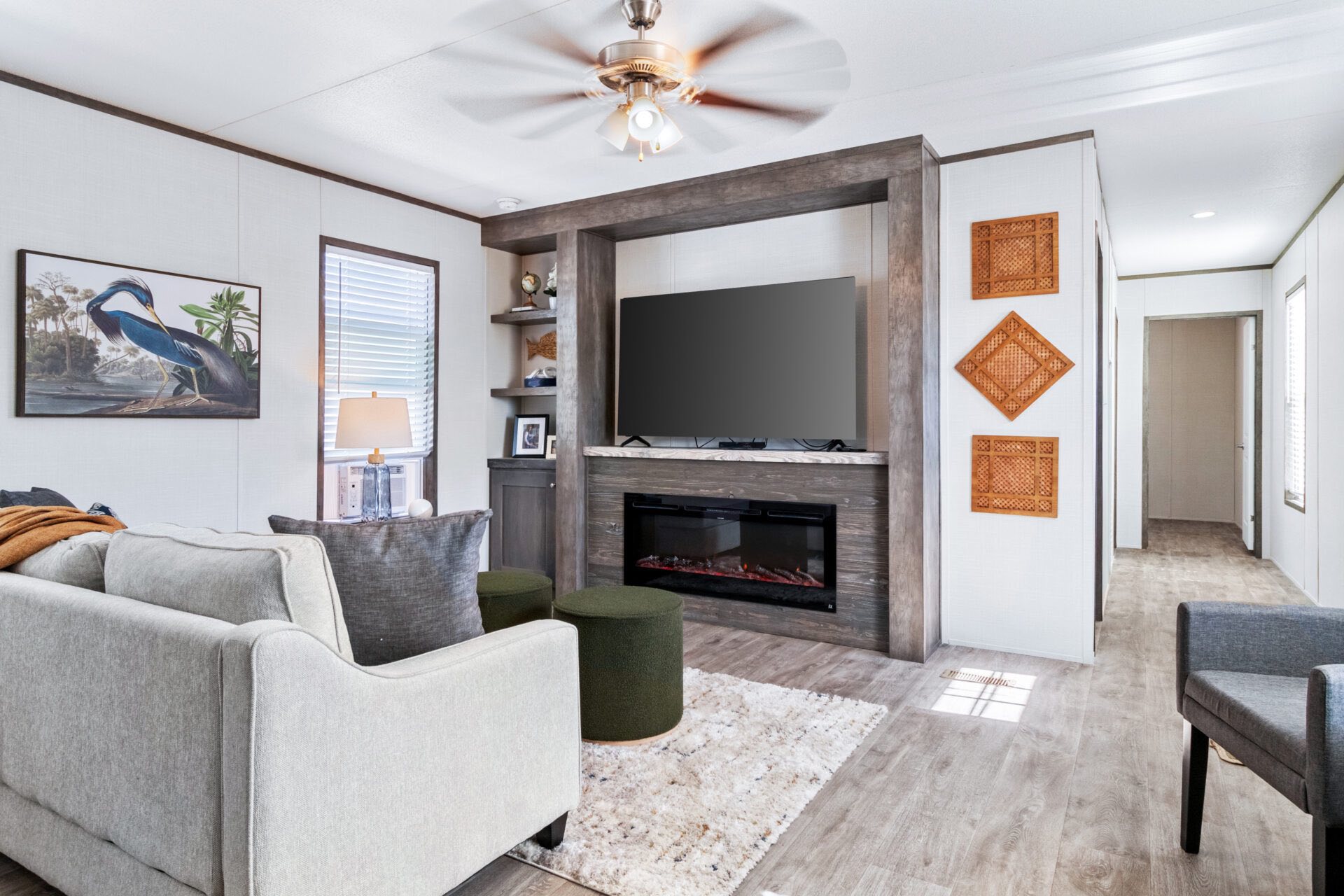 Modern living room with a plush gray sofa, a flat-screen TV on a wooden accent wall, and a cozy electric fireplace. A ceiling fan adds warmth.