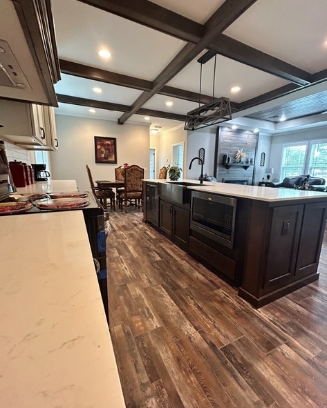 Spacious kitchen with dark wood floors, large island, and white countertops. Elegant dining area in the background, cozy living room to the right.