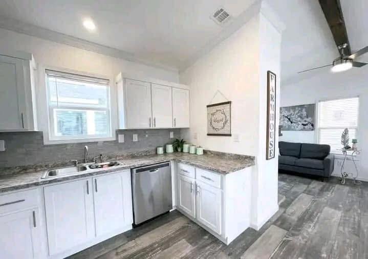 Bright kitchen with white cabinets, gray granite countertops, and stainless steel appliances. Sign reading "Farmhouse" and cozy living area visible. Calm, welcoming tone.