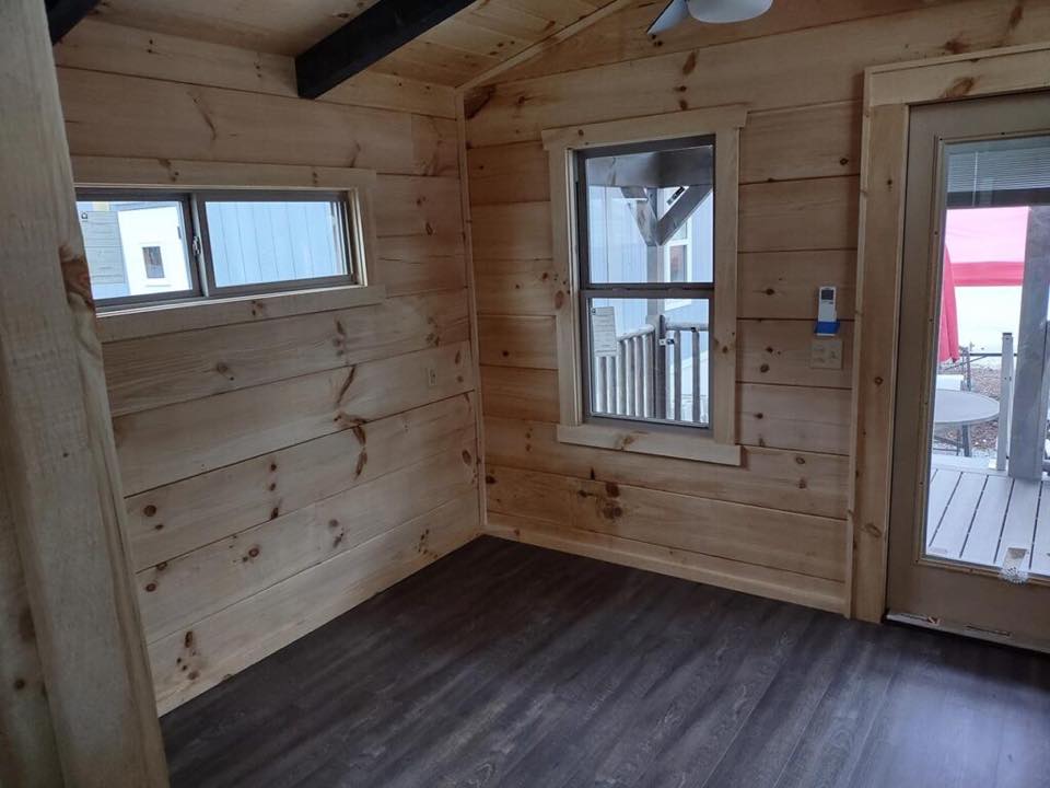 Cozy wooden room with light pine walls and dark wood flooring. A large window and glass door reveal an outdoor patio, creating a warm, inviting atmosphere.