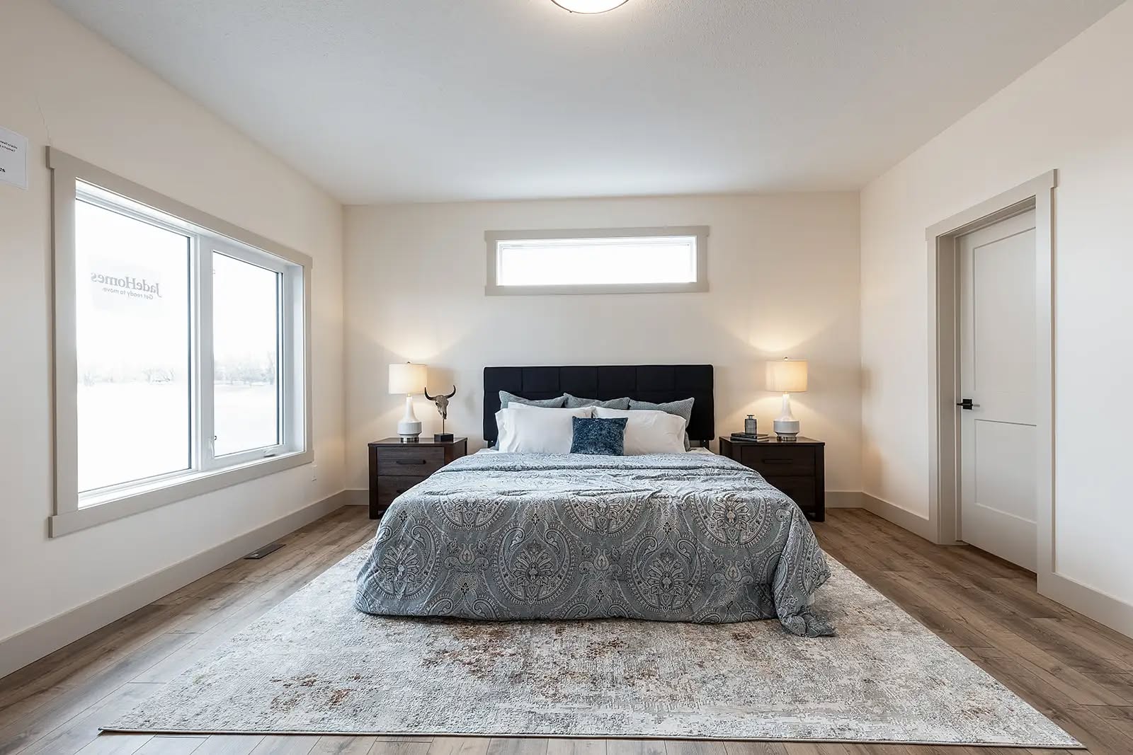 A cozy bedroom with a neatly made bed featuring a patterned gray comforter, white and blue pillows, flanked by two wooden nightstands with lamps. Natural light filters through a wide window, enhancing the calm and inviting atmosphere.
