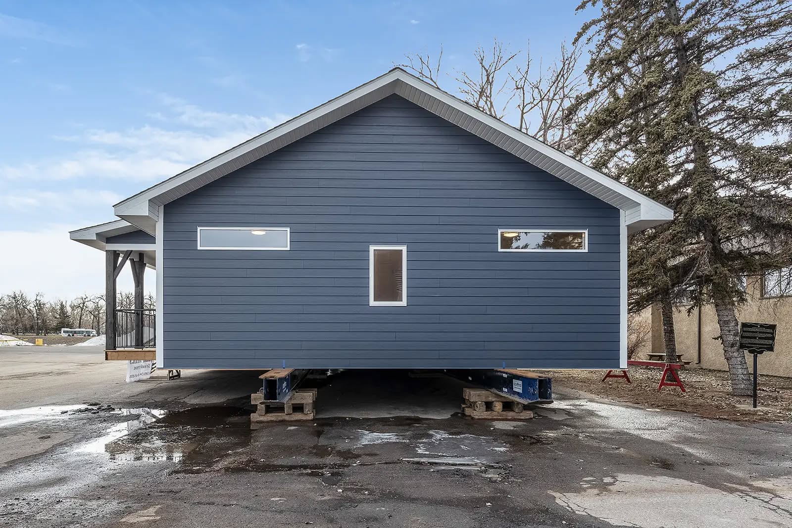 A blue modular house on wooden pallets, elevated off the ground, sits in a parking lot. Overcast sky, trees, and bare ground surround the structure.