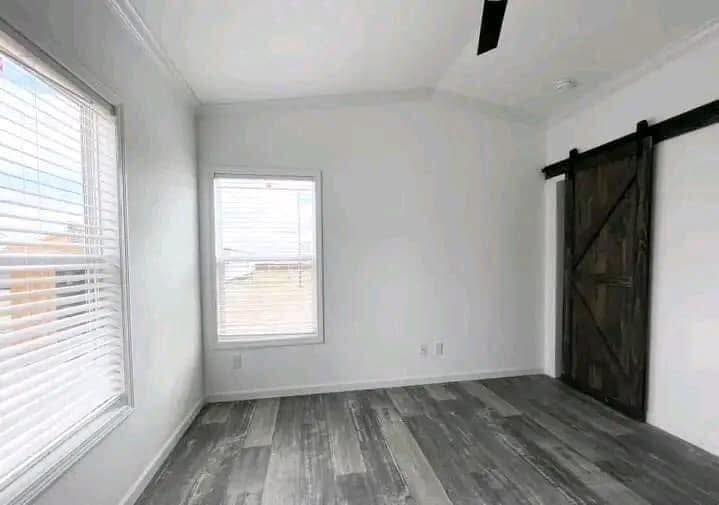 Bright, empty room with gray wood flooring, white walls, and a window with blinds. A dark wooden sliding barn door adds rustic charm.