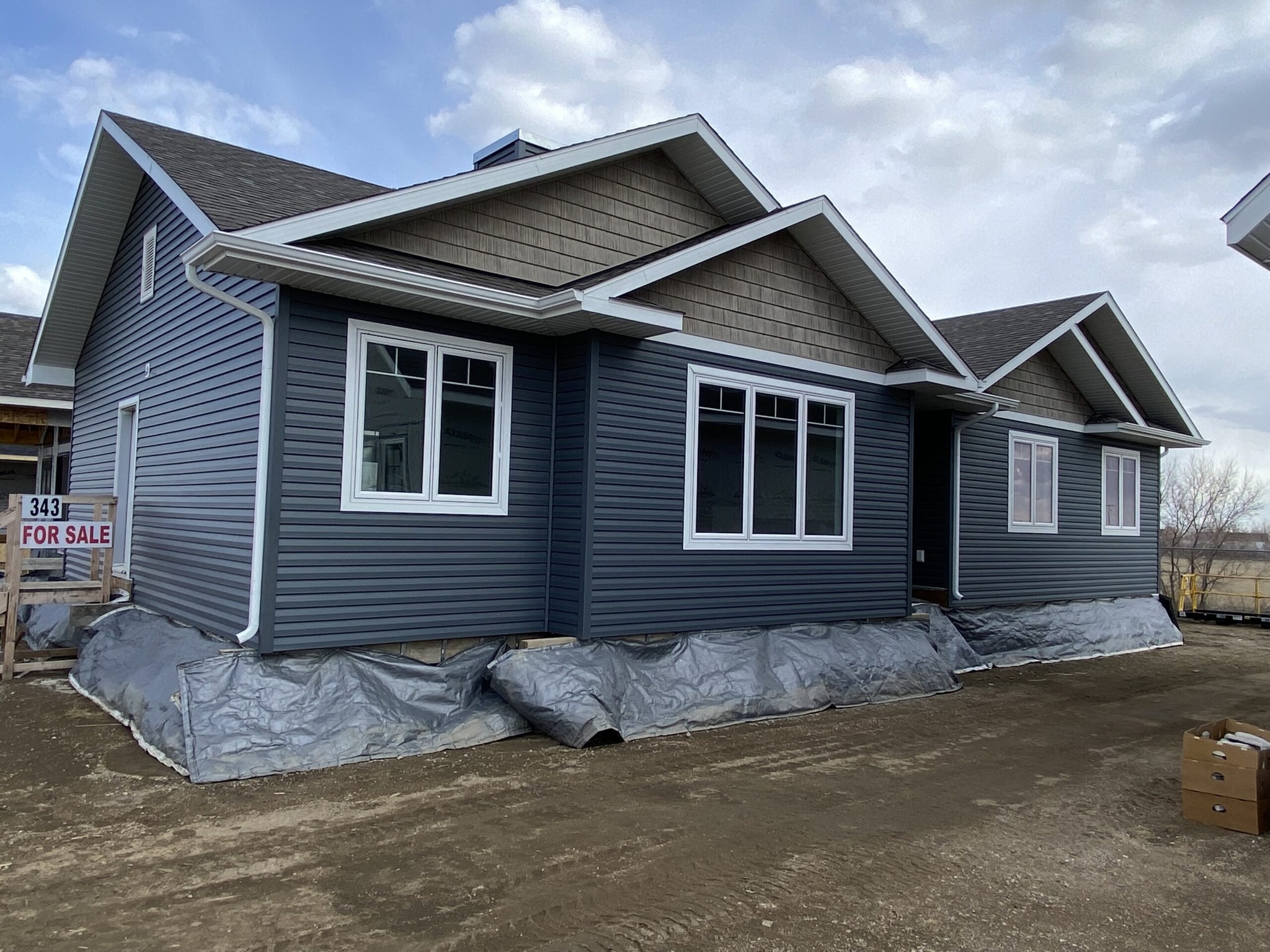 Newly constructed house with dark blue siding and white-trimmed windows. A "For Sale" sign is visible. Overcast skies set a muted, calm tone.
