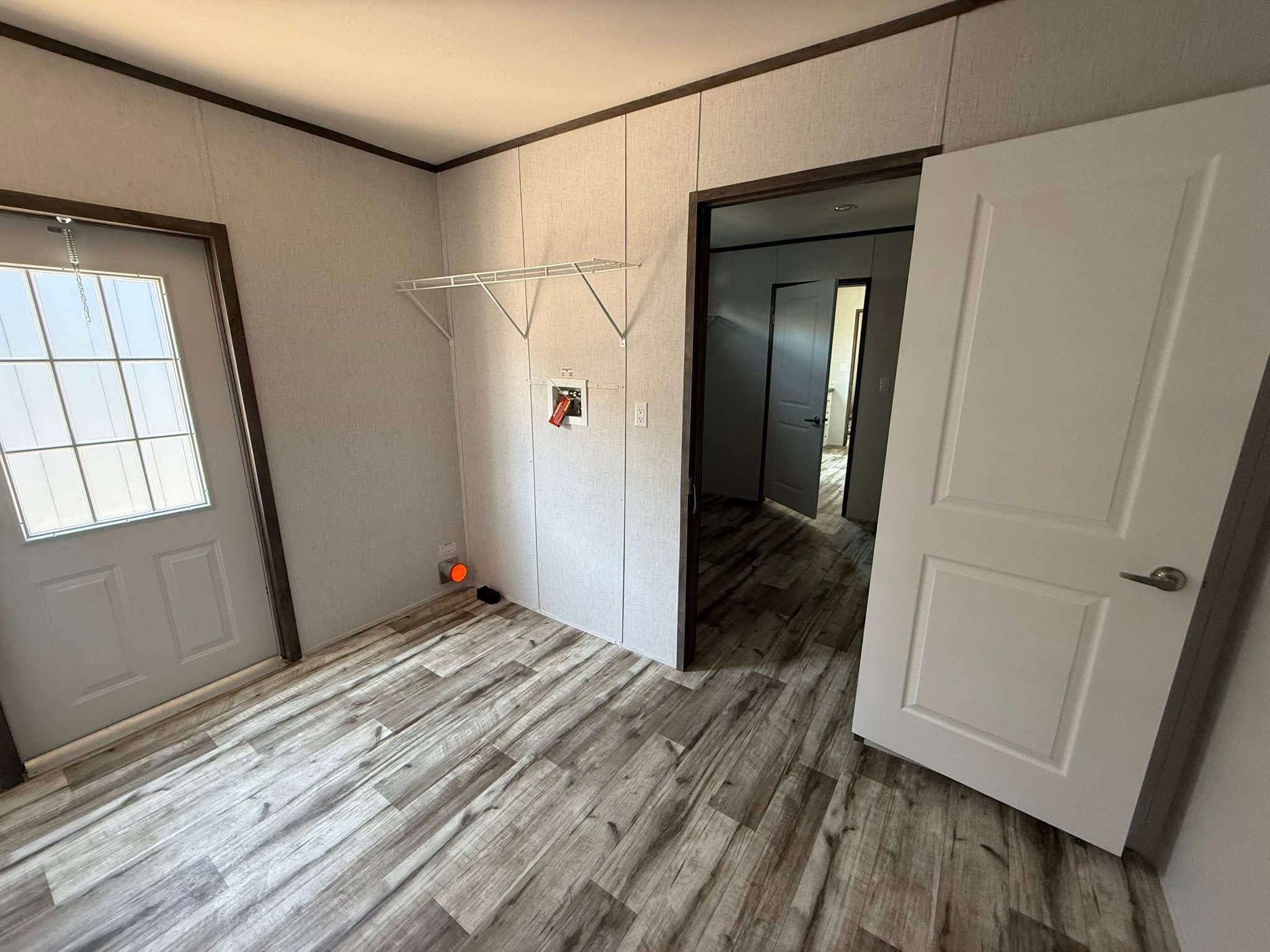 Bright room with light wood flooring, open door, and metal shelf. Wall has a control panel. Back door has a window. Calm, minimalistic space.