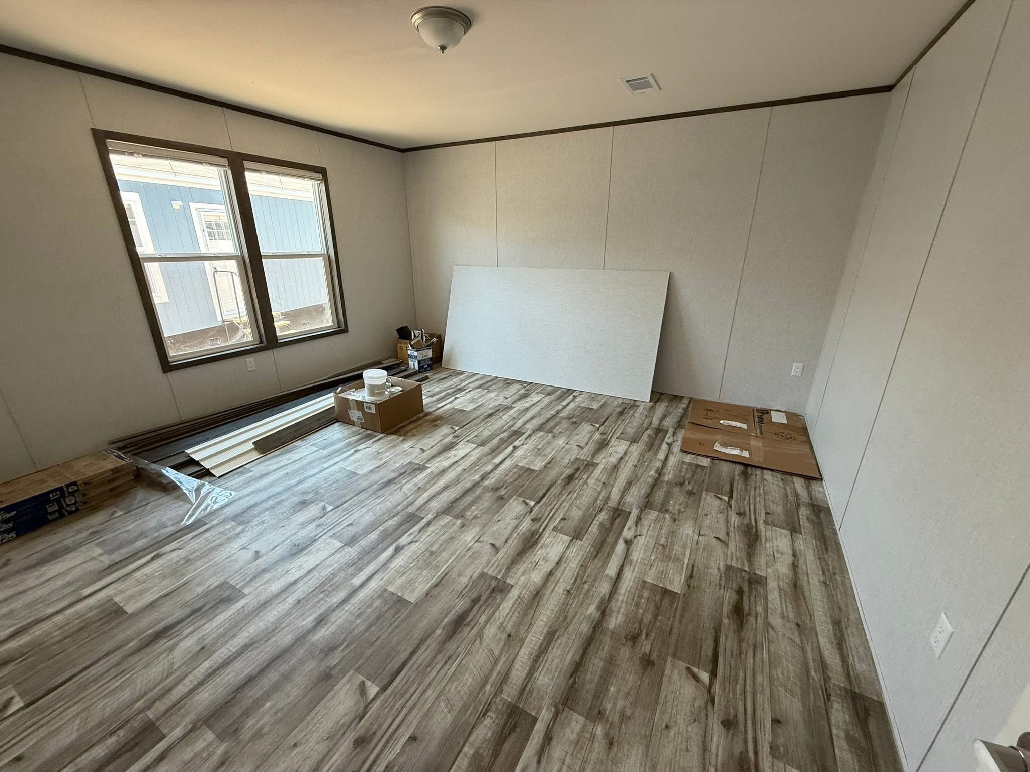 Spacious empty room with wooden floors, beige walls, and a small window on the left. Boxes and tools are scattered, indicating ongoing work.