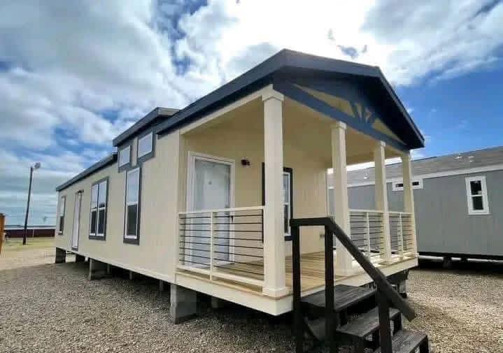 Small beige manufactured home with a covered porch and black railing. The sky is partly cloudy, creating a bright and open atmosphere.