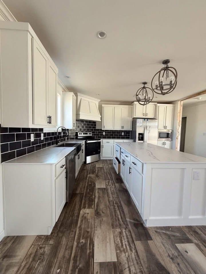 Modern kitchen with white cabinets, dark tiled backsplash, and wooden floor. Features large island, stainless steel appliances, and circular pendant lights.