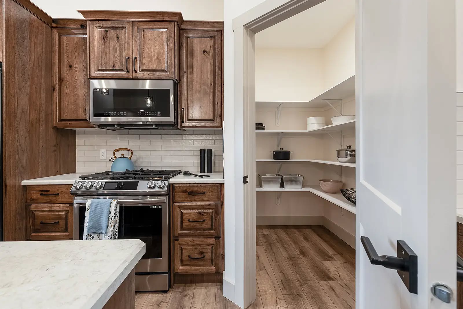 Modern kitchen with dark wood cabinets, a stainless steel oven, and a kettle on the stove. Open door reveals a tidy pantry with white shelves.