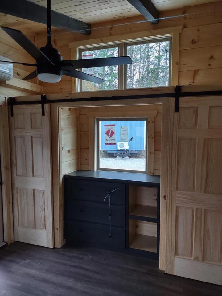 Interior of a wooden cabin with light wood walls, a dark ceiling fan, two sliding doors, a small window, and a black cabinet below. Cozy and rustic tone.